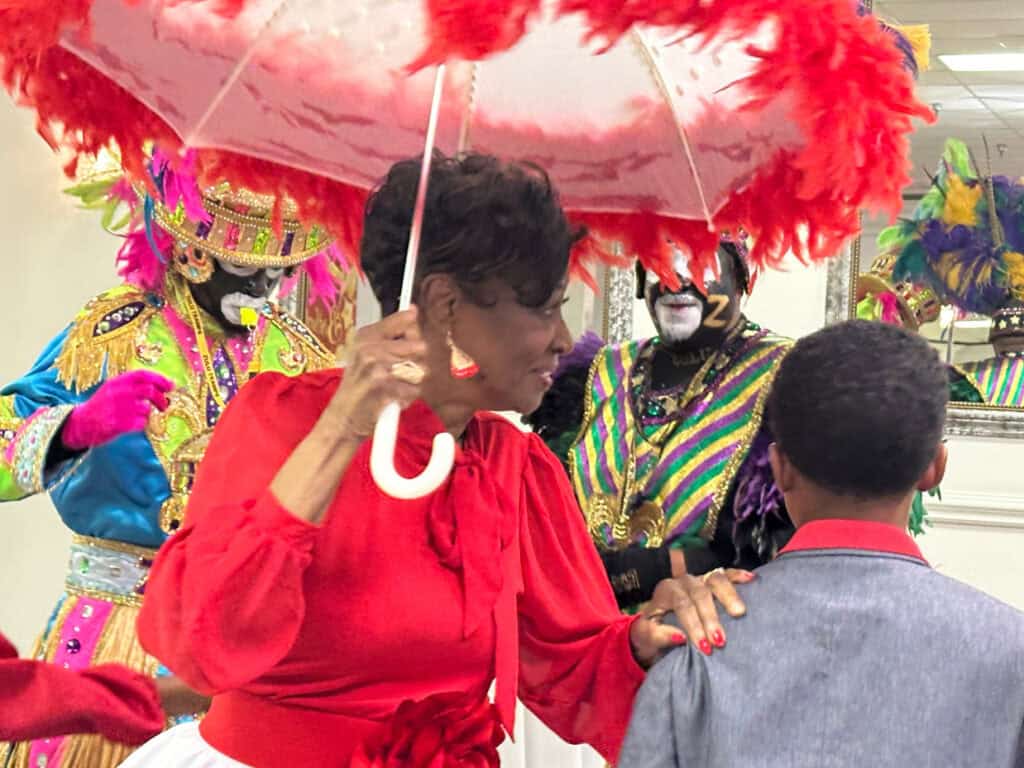 A woman in a red dress holds a decorative umbrella, accompanied by colorful performers in costumes and a boy in a gray suit.