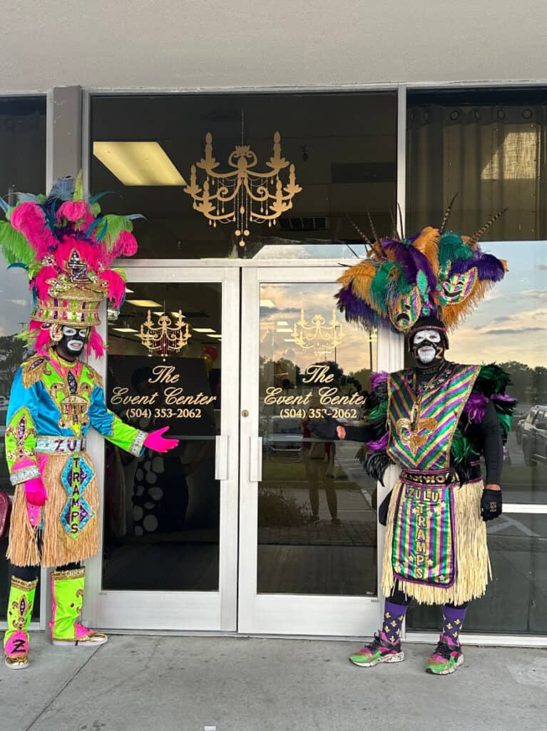 Two people dressed in vibrant, colorful Mardi Gras costumes stand in front of the double doors of The Event Center.