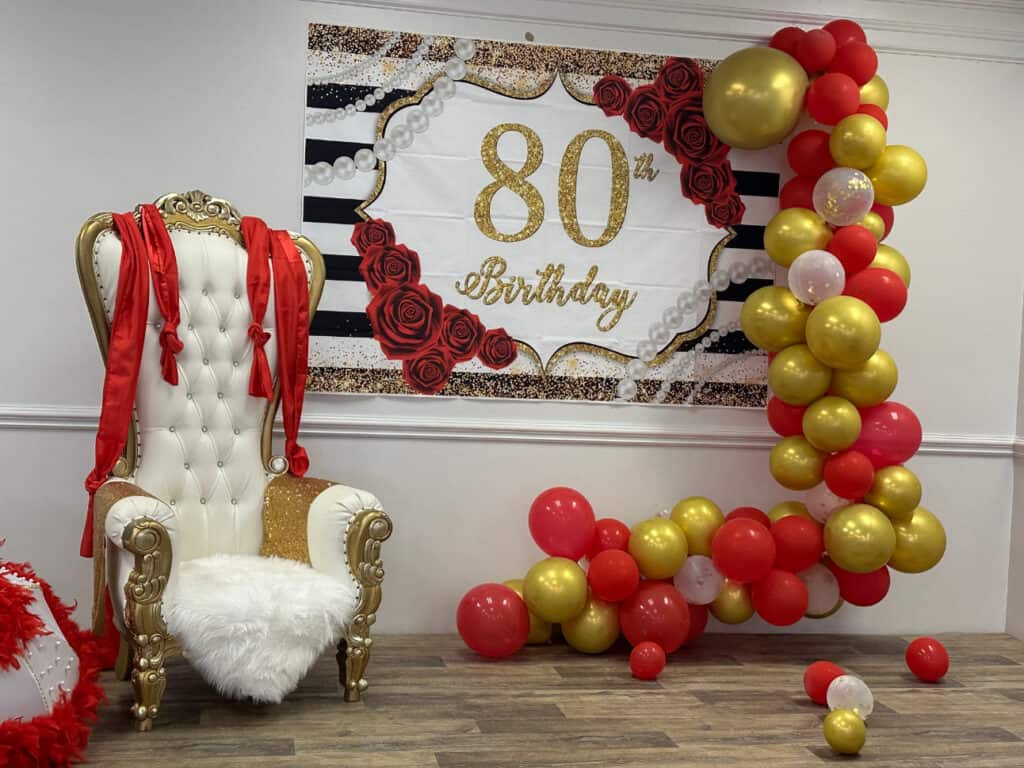 A chair with red ribbons is placed next to a banner that reads "80th Birthday" and a balloon arrangement in shades of red, white, and gold. The setup appears in a decorated indoor space.