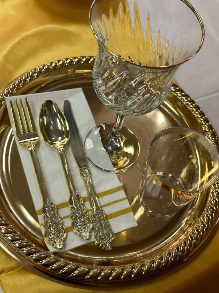 A gold-rimmed dinner plate with an ornate fork, spoon, knife, and napkin, alongside a decorative glass and a clear tumbler placed on a gold tablecloth.