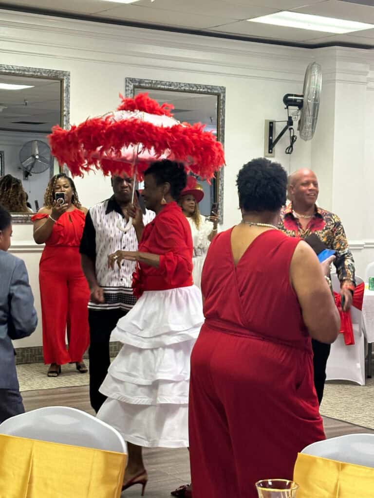 People dressed in vibrant red and white outfits participate in a festive indoor gathering. One person carries a large red feathered umbrella. Others socialize and take photos. .