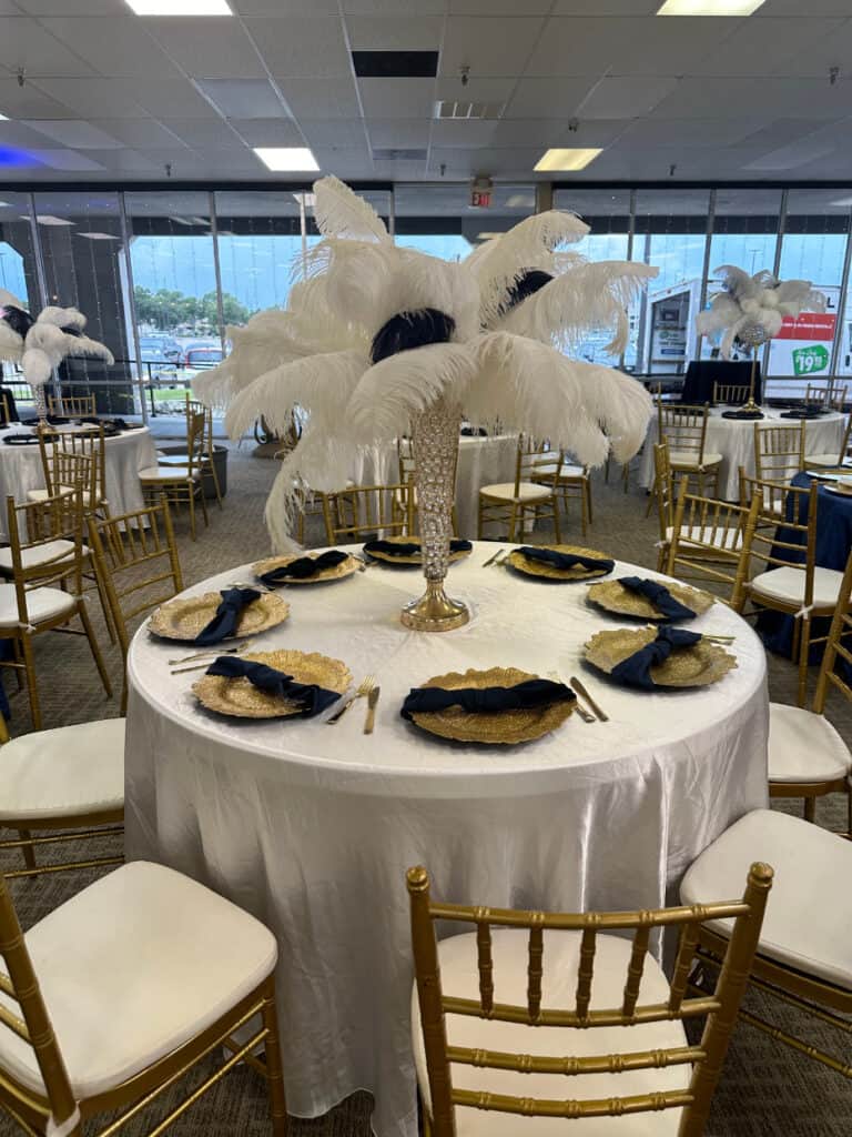 A round table with a white tablecloth, gold chairs, and a centerpiece of white feathers. Gold placemats, black napkins, and matching table settings are arranged on the table. Other similar tables are visible.