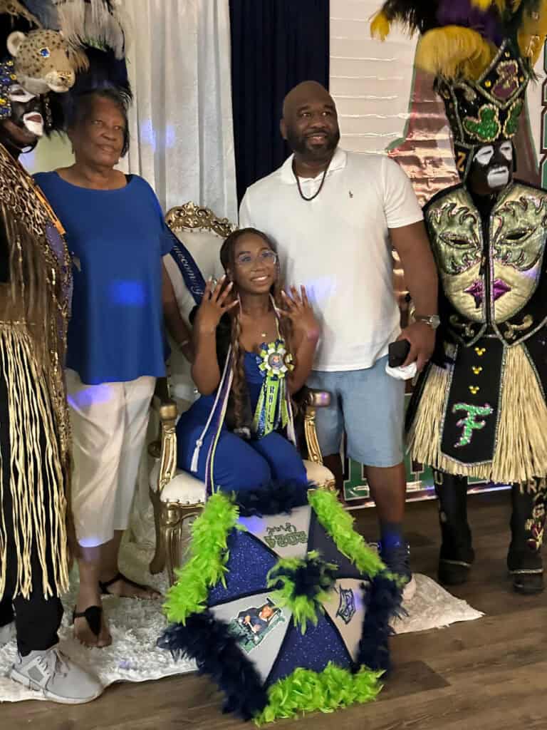 A woman sits on a throne-style chair, adorned in festive clothing and jewelry, flanked by people in elaborate costumes and two casually dressed individuals standing beside her.