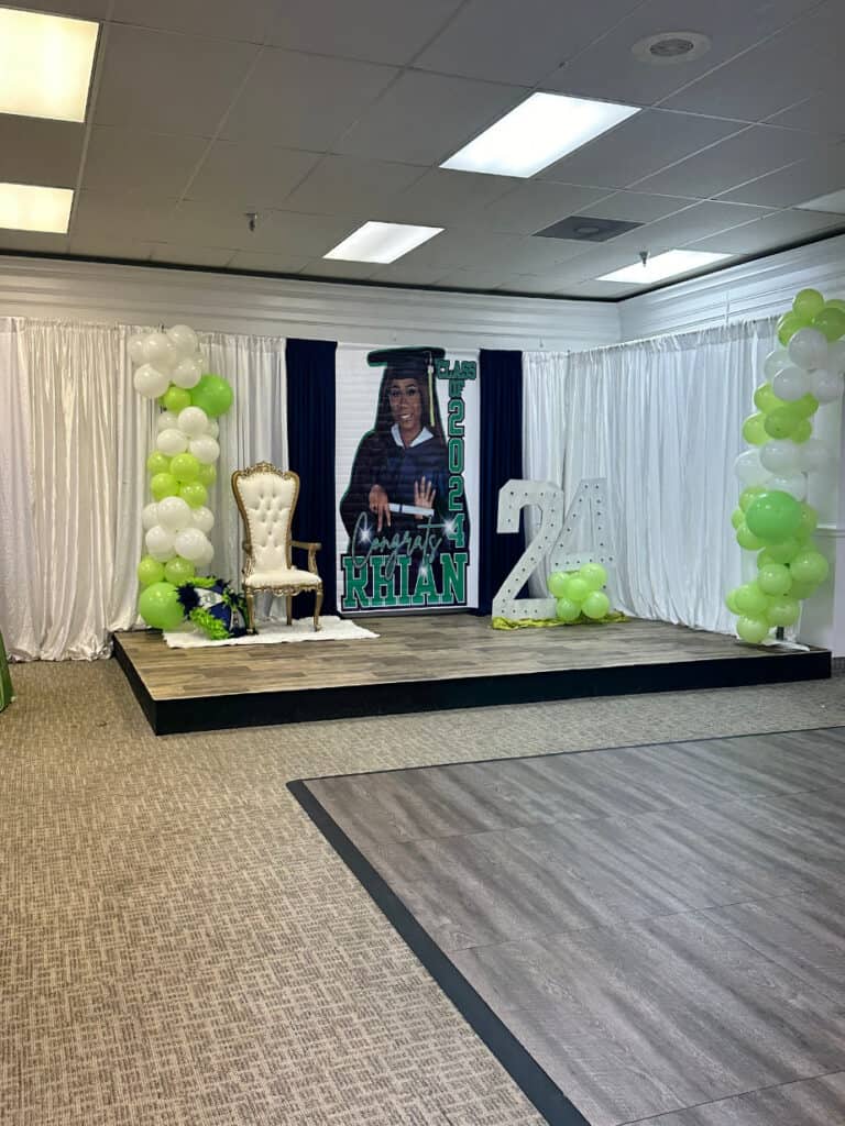 A decorated event stage featuring a large poster of a graduate with "Class of 2024" and the name "Kiran", a white and gold chair, and green and white balloons forming arches and the number "24".