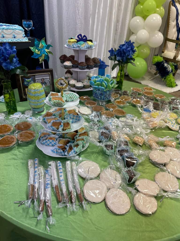 A table with a green tablecloth is filled with an assortment of baked goods, including cupcakes, cookies, and muffins, arranged around a cake stand and decorative items.