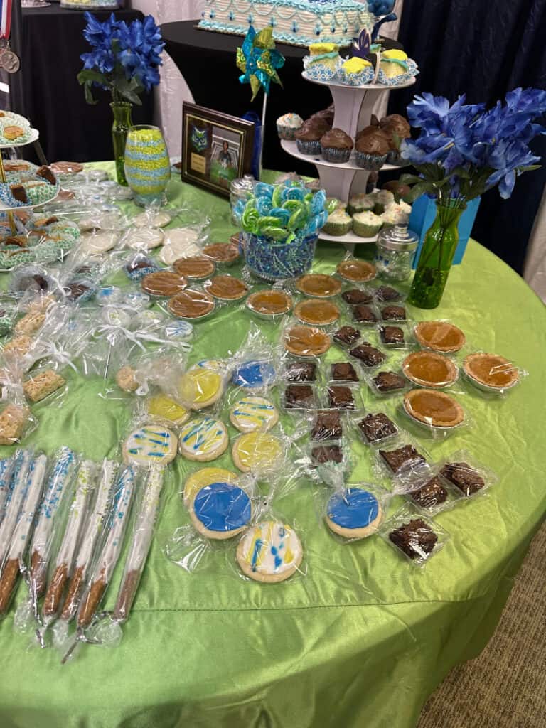 A table adorned with a variety of individually wrapped cookies, brownies, cake pops, and pastries. Vases with blue flowers and a decorative pinwheel accent the display.