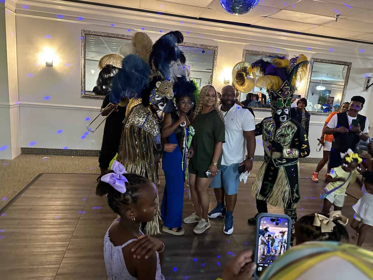 A group of people poses for a photo in a decorated room, some wearing ornate costumes with feathers and masks. Others, casually dressed, stand in the middle. A child in the foreground takes a picture.