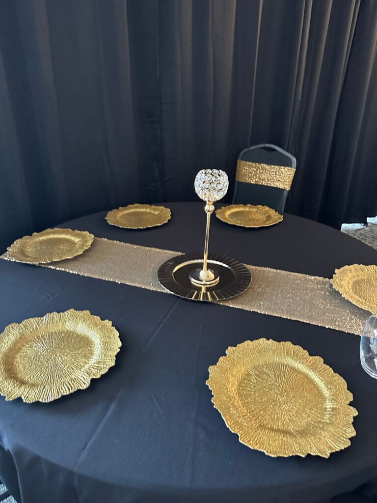 A round table set with gold-rimmed plates, a tall central candle holder with a spherical ornament, a black tablecloth, and a gold sequined runner against a black backdrop.