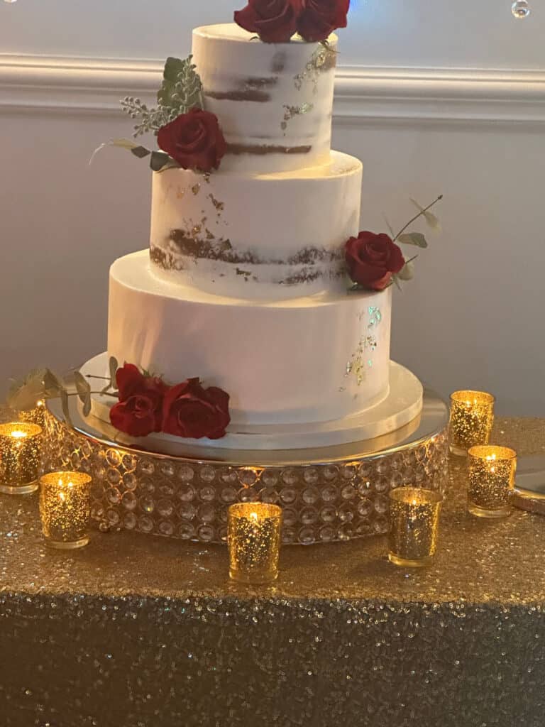 A three-tiered white cake decorated with red roses and surrounded by lit candles on a glittering tablecloth.