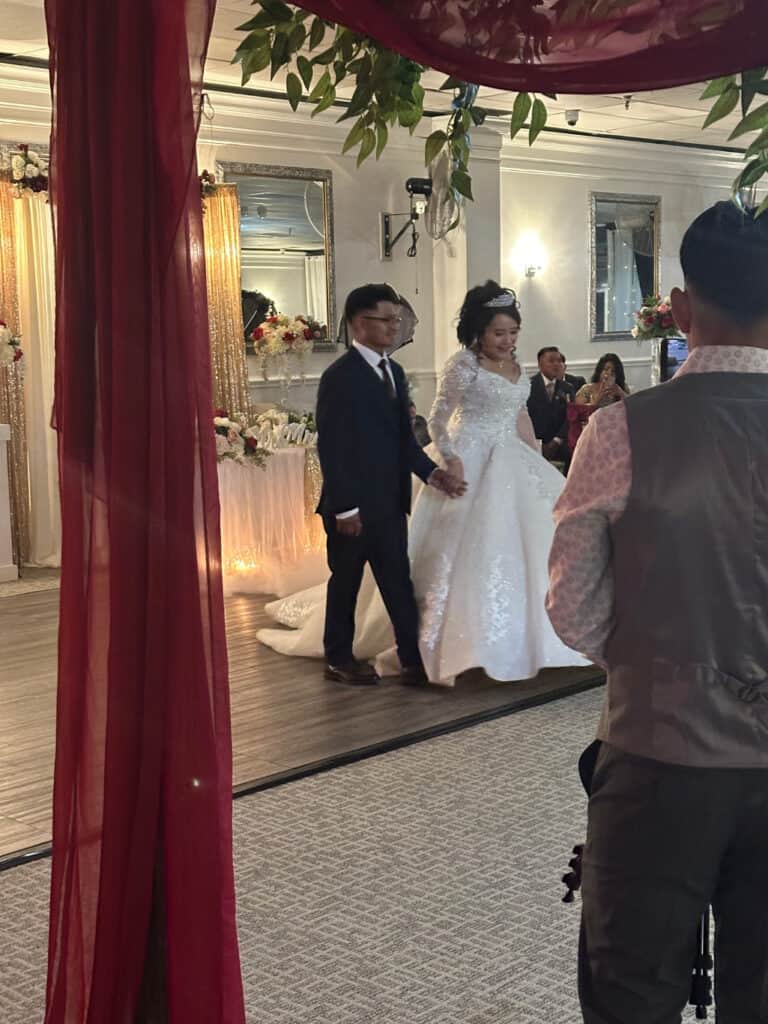 A bride and groom hold hands while walking at a wedding ceremony. The bride wears a white dress with a train, and the groom is in a dark suit. Other guests are present in the background.