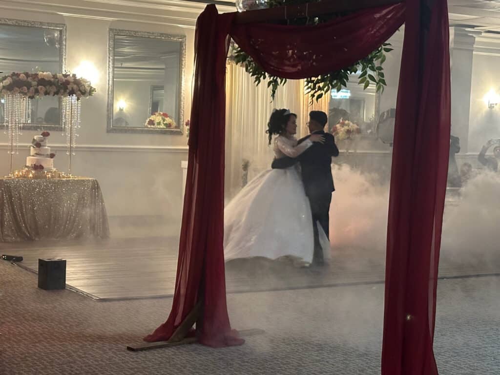 A bride and groom share their first dance on a foggy dance floor, framed by red drapery, with a wedding cake and floral decorations visible in the background.