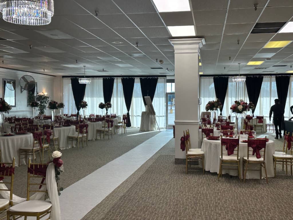 A banquet hall is set for an event with round tables covered in white tablecloths and red napkins. Floral centerpieces adorn the tables, and a white aisle runs through the center.