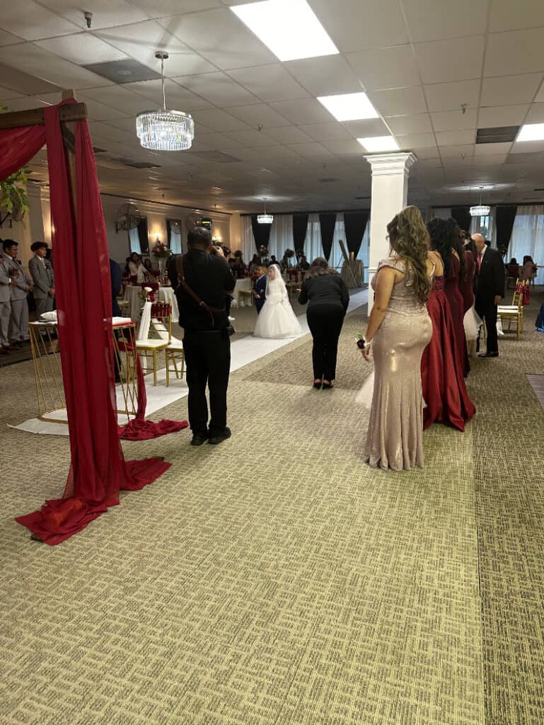 A bride in a wedding gown walks down the aisle in a decorated indoor venue, accompanied by guests and photographers. Groomsmen and bridesmaids stand on either side, and chairs are arranged for attendees.