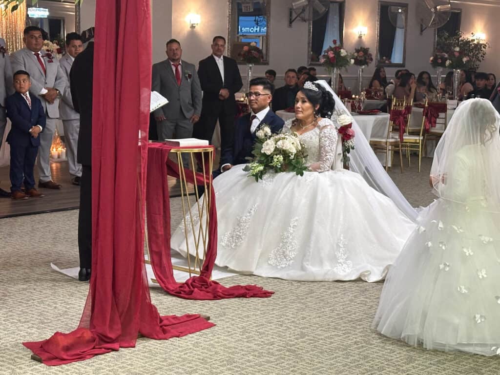 A bride and groom kneel beside a priest during a wedding ceremony in a decorated hall, with guests and a flower girl looking on.